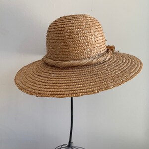 Pu&ograve; includere: Un cappello di paglia a tesa larga con una fascia intrecciata marrone e un piccolo fiocco su un lato. Il cappello &egrave; esposto su un supporto nero.