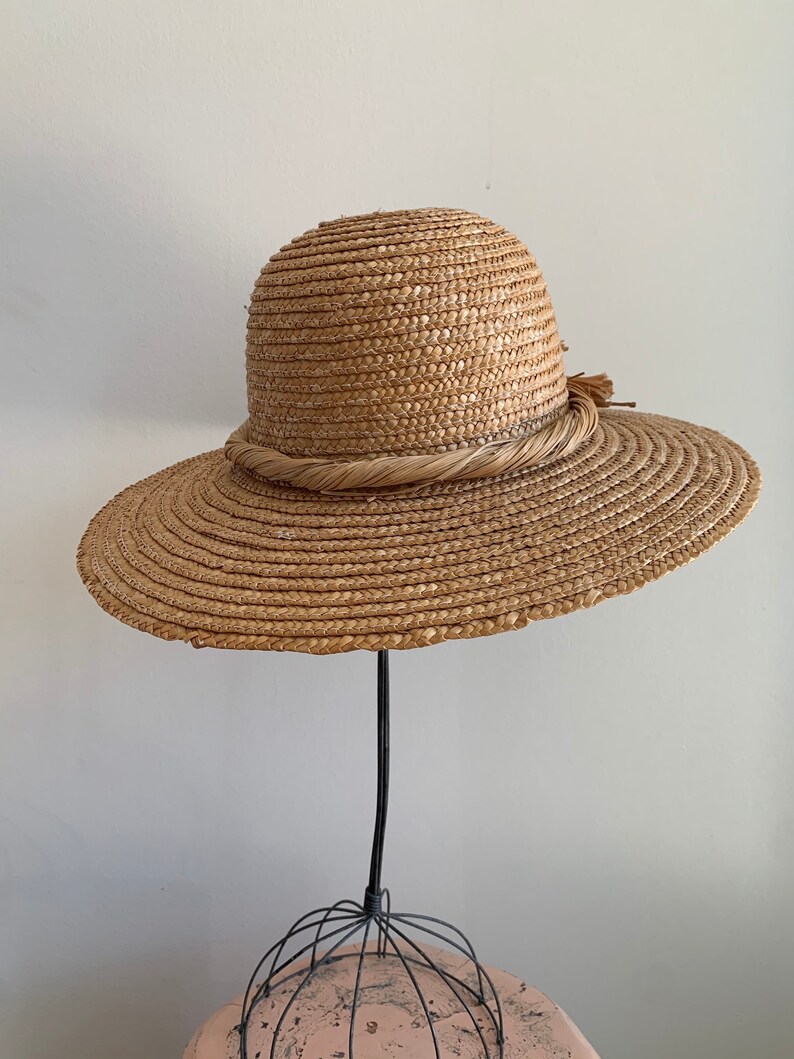 Pu&ograve; includere: Un cappello di paglia a tesa larga con una fascia intrecciata marrone e un piccolo fiocco su un lato. Il cappello &egrave; esposto su un supporto nero.
