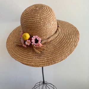 Pu&ograve; includere: Un cappello di paglia da sole con un'ampia tesa e un nastro marrone. Il cappello &egrave; decorato con fiori rosa e un limone giallo.
