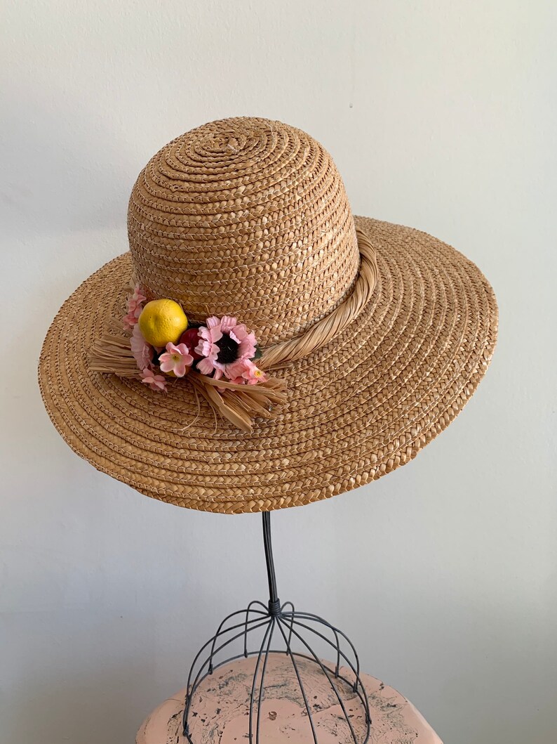 Pu&ograve; includere: Un cappello di paglia da sole con un'ampia tesa e un nastro marrone. Il cappello &egrave; decorato con fiori rosa e un limone giallo.