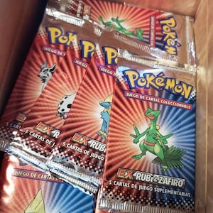 May include: A box full of Pokemon trading card game booster packs. The packs are red, blue, and silver with images of Pokemon characters. The text on the packs reads "Pokemon Juego de Cartas Coleccionable" and "Ex Rubi & Zafiro 5 Cartas de Juego Supplementarias".