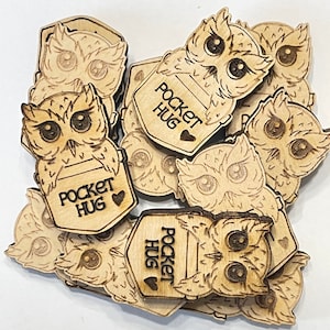 Könnte beinhalten: Ein Stapel hölzerner Eulenschmuckstücke mit dem Text "Pocket Hug" und einem kleinen Herz auf jedem Stück.