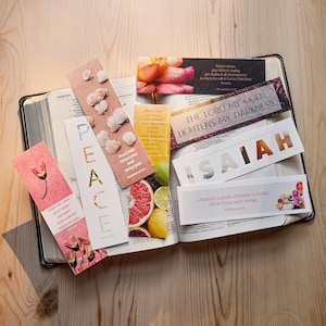 May include: A collection of rectangular paper bookmarks featuring various designs, including seashells, citrus fruits, flowers, and inspirational text such as PEACE, ISAIAH, and THE LORD MY GOD LIGHTENS MY DARKNESS, resting on an open book.