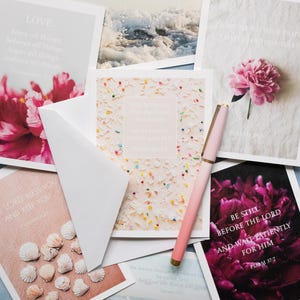 May include: A collection of rectangular greeting cards featuring floral, ocean, and seashell imagery with printed scripture verses, accompanied by a light pink pen with gold accents. The cards display various religious texts and colorful designs.