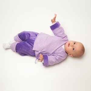 Baby trägt eine lilafarbene FIORINO Wickeljacke aus gepunktetem Stoff mit langen Ärmeln, breiten violetten Manschetten und seitlichen Bindeschleifen