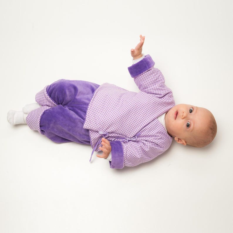 Baby trägt eine lilafarbene FIORINO Wickeljacke aus gepunktetem Stoff mit langen Ärmeln, breiten violetten Manschetten und seitlichen Bindeschleifen