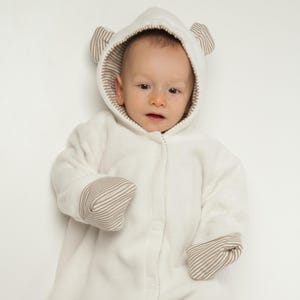 May include: A cream-colored, long-sleeved baby bunting with an attached hood featuring small bear ears. The interior of the hood and the fold-over mitten cuffs are lined with a brown and white striped fabric. The garment has a front snap closure.