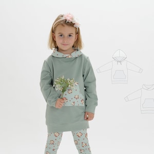 Könnte beinhalten: Ein junges Mädchen trägt einen hellgrünen Kapuzenpullover mit einer Tasche mit Blumendruck. Der Pullover hat eine kontrastierende Blümchen-Futterung um die Kapuze. Das Mädchen hält einen kleinen Strauß weißer Blumen.