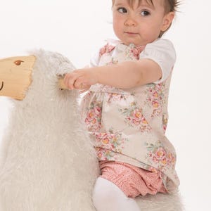 Kleinkind sitzt auf einem weißen Kuschelschaf und trägt ein geblümtes LIPSIA Trägerkleid über einem weißen Shirt mit langen Ärmeln.