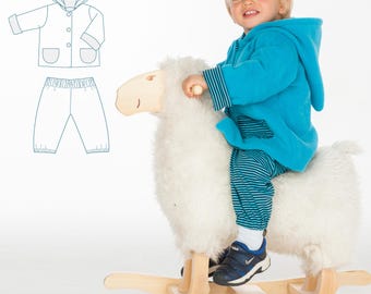 Könnte beinhalten: Ein kleines Kind in einer blauen Kapuzenjacke und gestreiften Hosen reitet auf einem weißen Schaukelpferd. Das Kind lächelt und schaut nach links. Im Hintergrund ist ein Schnittmuster für eine Kapuzenjacke und eine Hose zu sehen.