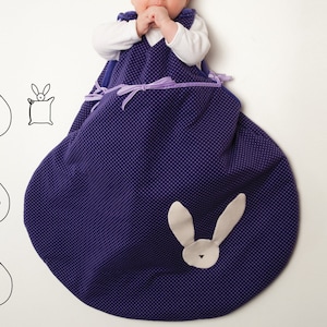 May include: A purple and white baby sleeping bag with a bunny design. The sleeping bag has a zip closure and a hood. The sleeping bag is made of soft, comfortable fabric.