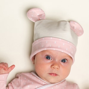 May include: A baby wearing a pink and white hat with bear ears and a pink and white polka dot onesie.