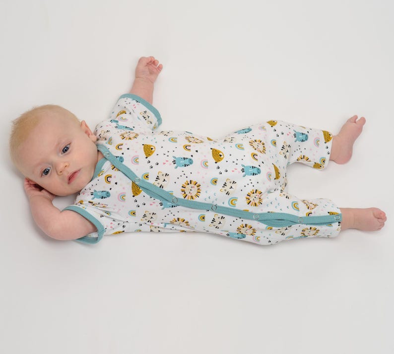 Baby liegt in einem weißen kurzärmeligen LEON Sommerstrampler mit Tier- und Regenbogenmuster, türkiser Einfassung und diagonaler Druckknopfleiste