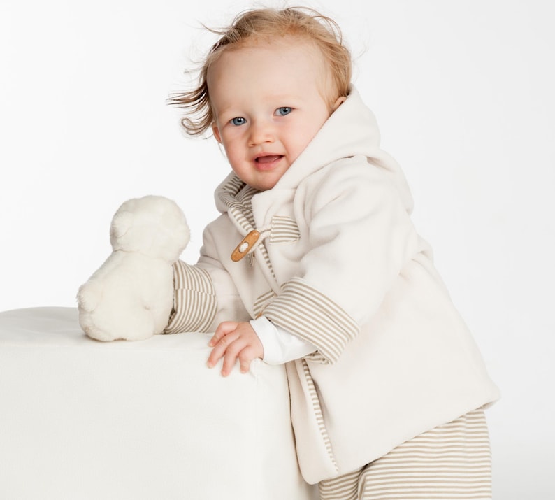 Kleinkind trägt eine cremefarbene BRIO Kapuzenjacke mit Knöpfen und die gestreifte LUCCA Wendehose, komplettes Baby-Schnittmuster-Set.