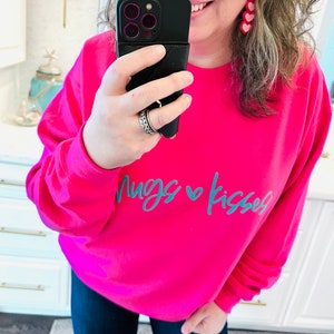 May include: A pink sweatshirt with the text "Hugs & Kisses" in teal blue lettering. The sweatshirt has long sleeves and a crew neck.