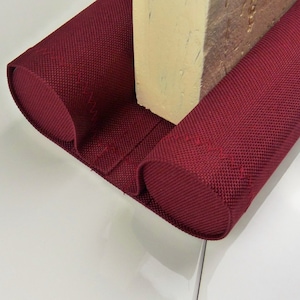 Op de afbeelding: Een bordeauxrode tochtstrip voor de deur van een geweven stof. De tochtstrip is om een lichtgekleurde houten paal gewikkeld. De stof is aan de uiteinden omgeslagen om een afgeronde vorm te creëren.