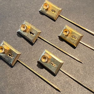May include: Five gold-toned keyhole pins with a small knob on the top. The pins are arranged in a row on a gray surface.