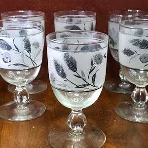 May include: Set of six vintage clear glass goblets. Each goblet features a frosted upper portion with a floral and wheat design in gray and black. The goblets have a round base and a decorative stem.