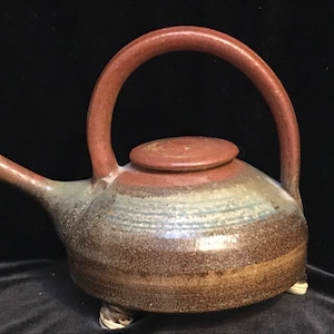 May include: A handmade ceramic teapot with a brown handle and lid. The body of the teapot features bands of brown, blue, and tan colors. The teapot has a spout and sits on three small feet.