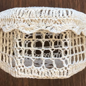 May include: A handmade, off-white crochet basket with a scalloped edge. The basket has a cylindrical shape with an open, lattice-like design. The crochet work appears to be made of a natural fiber, giving it a rustic and textured appearance. The basket is likely used for storage or decoration.
