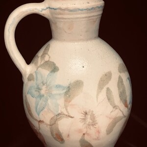 May include: White ceramic pitcher with a rounded body and a curved handle. The pitcher is decorated with hand-painted blue and pink floral designs and green leaves. A blue wavy line accents the rim.