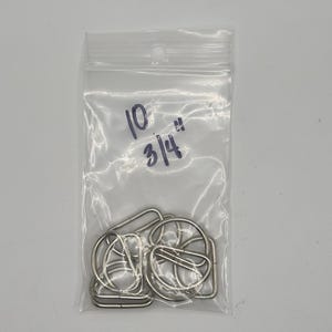 May include: A clear plastic resealable bag containing several silver-colored metal D-rings. The bag has the text 10 and 3/4" written in blue ink on the front, indicating the quantity and size of the hardware pieces contained inside the package.
