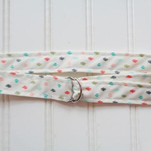 May include: A white fabric strap with a silver D-ring. The strap has a repeating pattern of small, colourful geometric shapes.