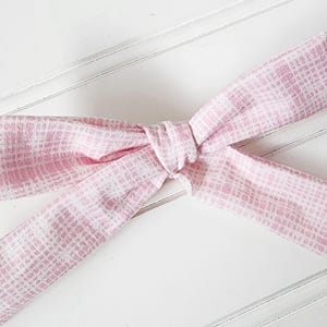 May include: A pink and white patterned fabric bow, tied in the center. The bow is made of a soft material and is set against a white background.