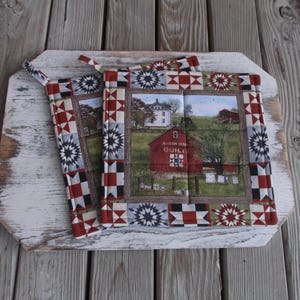 May include: Two red, white, and blue quilted pot holders with a farmhouse scene featuring a red barn with the text "Auld Mill Quilts" and a white house. The pot holders have a starburst pattern.