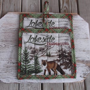 May include: Two green, red, and white plaid pot holders with a white background and the text "Lakeside Campground" in black. The pot holders feature a fox and pine trees in a winter scene.