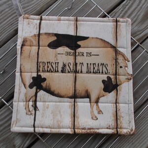 May include: A white quilted pot holder with a brown and black illustration of a pig. The text "DEALER IN FRESH & SALT MEATS." is printed on the pot holder.