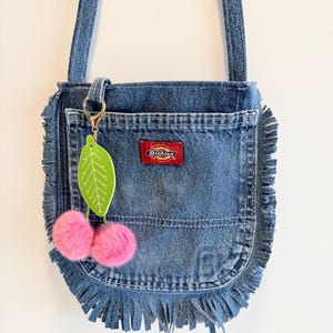May include: A blue denim shoulder bag with frayed edges and a long strap. The front features a rectangular pocket with a red Dickies brand label. A keychain with a green leaf charm and two fuzzy pink pom-poms is attached to the bag's hardware.