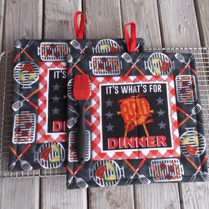 May include: Two red and black pot holders with a checkered pattern and a grill theme. The text on the pot holders reads "It's what's for BBQ Dinner".