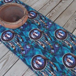 May include: A teal table runner with a repeating pattern of dreamcatchers with feathers. The dreamcatchers have a brown and white horse head in the centre. The table runner is on a wooden surface.