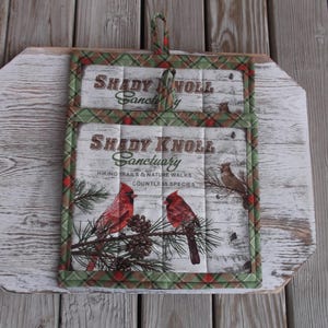 May include: Two red and green plaid pot holders with the text "Shady Knoll Sanctuary" and a picture of cardinals and pine branches.