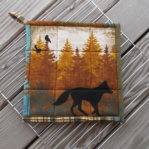 May include: A brown and green quilted pot holder with a black silhouette of a fox and birds in a forest scene.