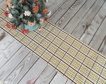 May include: A plaid table runner in shades of tan, green, and brown, with dark brown stripes. The runner is placed on a wooden surface, and a vase of flowers sits at the top. The flowers are a mix of red, white, and orange.