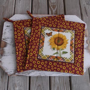 May include: Two burgundy pot holders with a sunflower and butterfly design. The pot holders have a repeating pattern of yellow sunflowers on a burgundy background. The centre features a sunflower and butterfly.