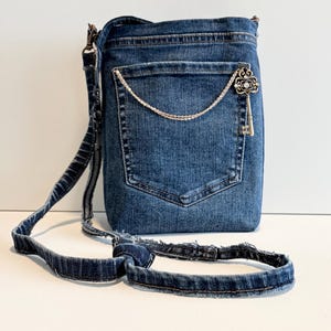 May include: A blue denim crossbody bag featuring a single external pocket with a decorative silver-toned chain and a vintage-style skeleton key charm. The bag includes a matching frayed denim strap with a knotted adjustment detail on the side.