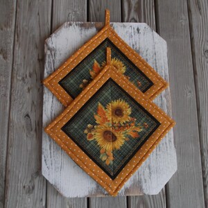 May include: Two quilted pot holders featuring a sunflower and autumn leaf design. The pot holders are yellow with white polka dots and have a dark green background.