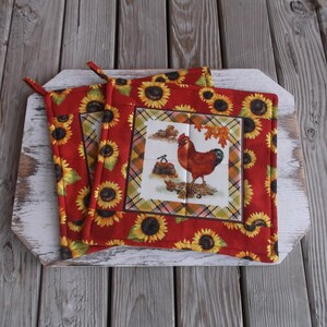 May include: Two red pot holders with a sunflower and plaid pattern. One pot holder features a rooster illustration.