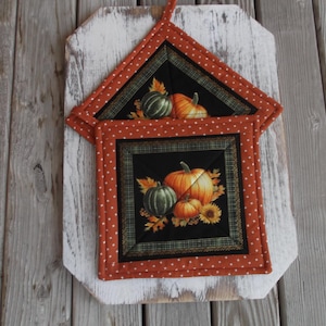 May include: Two orange and white quilted pot holders with a black background featuring a fall harvest theme with pumpkins and leaves.