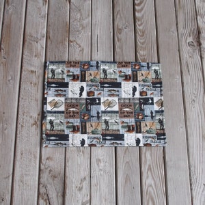 May include: A rectangular placemat with a fishing theme. The placemat is made of fabric and features a patchwork design with various images of fish, fishing rods, tackle boxes, and the text "Catch of the Day".