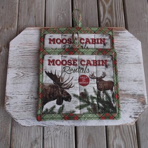 May include: Two green and red plaid pot holders with a moose and pine tree design. The pot holders have the text "Moose Cabin Rentals" and "Daily & Weekly" printed on them.