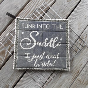 May include: A gray and white quilted hot pad with the text "Climb into the Saddle I just need to ride!" printed on it. The hot pad has a white border with a small black and white pattern.