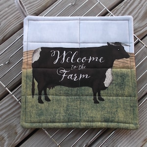 May include: A quilted pot holder with a black and white cow silhouette and the text "Welcome to the Farm". The background is a green and brown field.