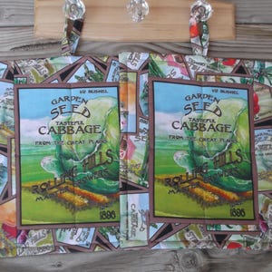 May include: A quilted fabric book cover featuring a vintage-style seed packet design with the words "Garden Seed Tasteful Cabbage" and a landscape illustration. The cover is decorated with floral and seed packet patterns.