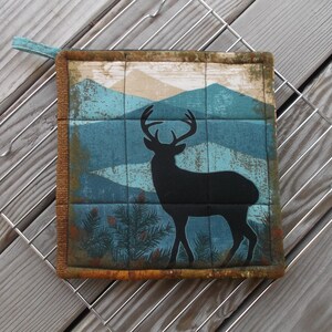 May include: A blue and brown pot holder with a silhouette of a deer in front of a mountain range.