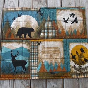 May include: A quilted placemat with a woodland theme. The placemat features a black bear, a deer, a cabin, and birds against a backdrop of trees, mountains, and a moon.