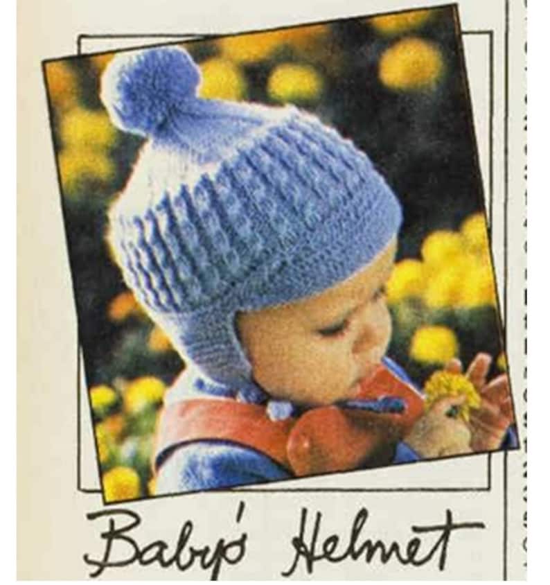 May include: A blue knitted baby helmet with a pom-pom on top. The helmet is worn by a baby who is looking down at a flower.